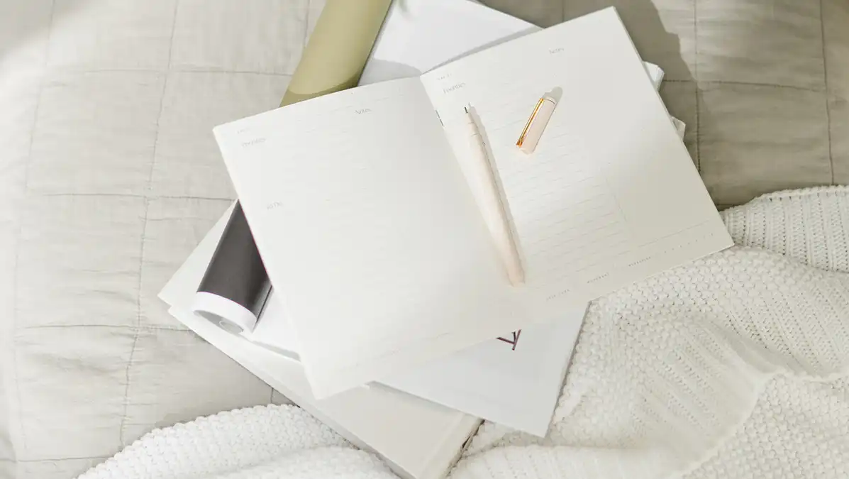 Een open notitieboek met pen op een bed, naast een kop koffie en groen blad – een uitnodiging om jouw ideale werkleven op te schrijven.