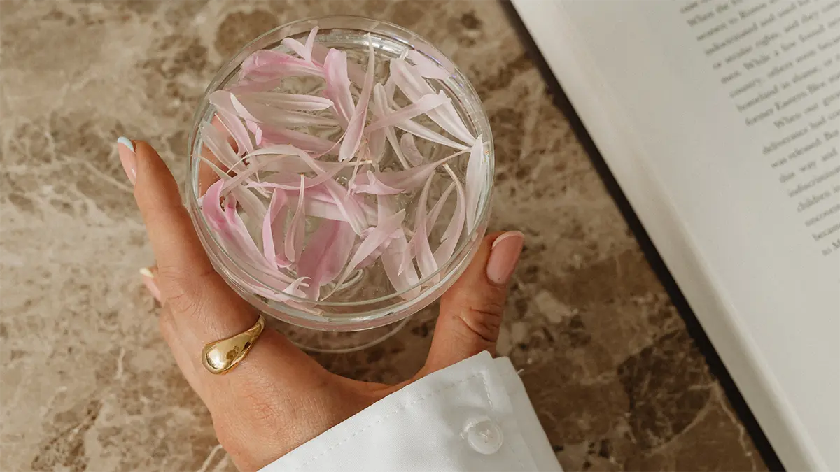 Vrouwelijk hand met glas water op een open boek – een rustmoment tussen de to-do’s, symbool voor vertraging en thuiskomen bij jezelf.