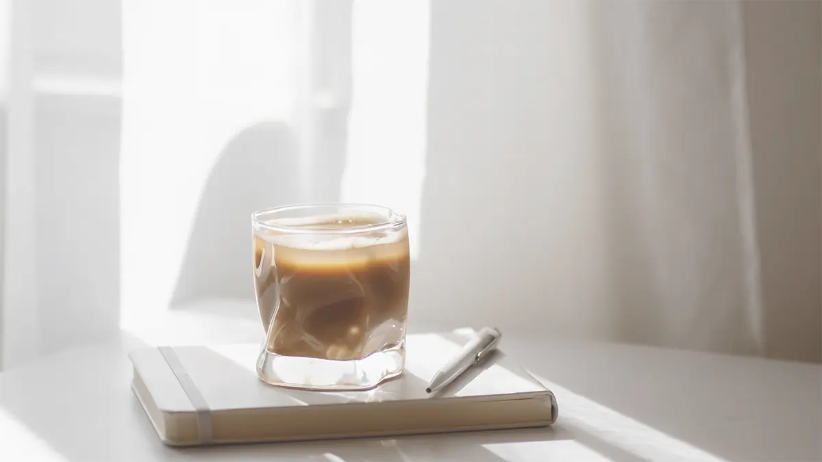 Glas koude koffie op een notitieboek met pen in zacht ochtendlicht – een moment van stilte, journaling en reflectie.