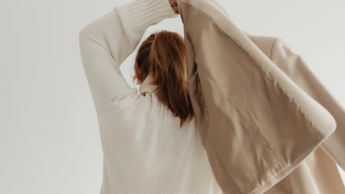 Vrouw in crèmekleurige trui houdt een beige jas vast. Twijfelmoment. Kiezen wat klopt. Lichaamssignalen, werk, keuzes